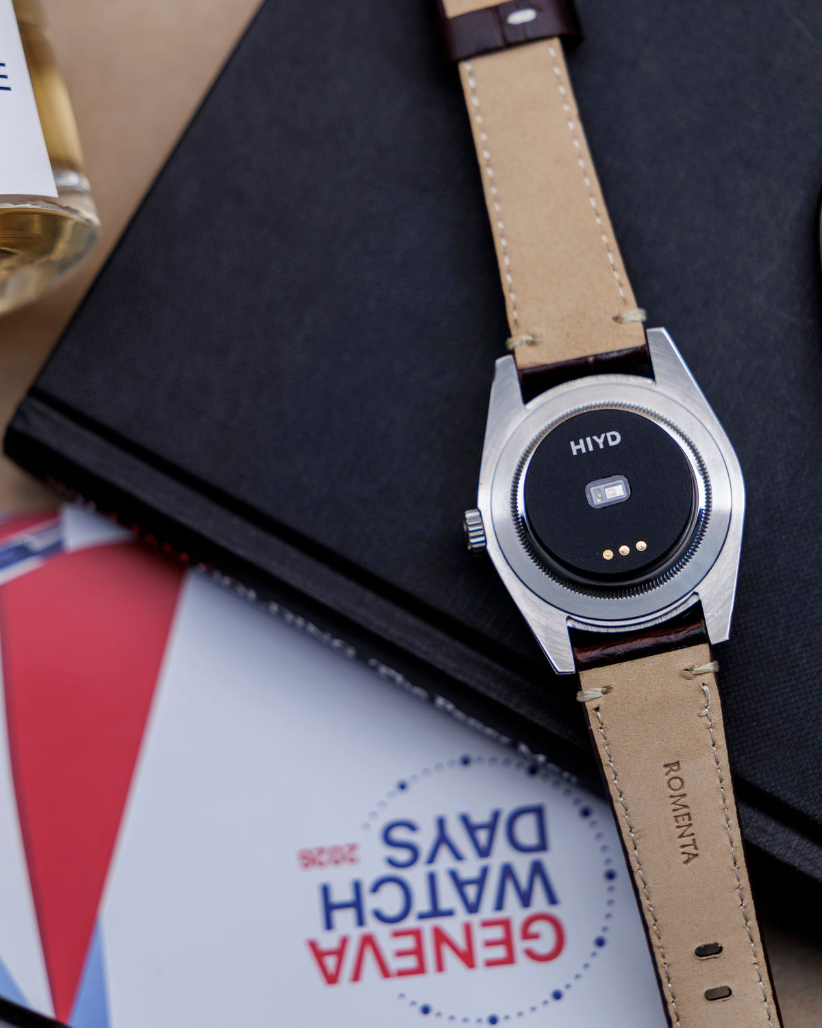 A steel watch with HIYD fitted, on a black notebook beside a Geneva Watch Days card