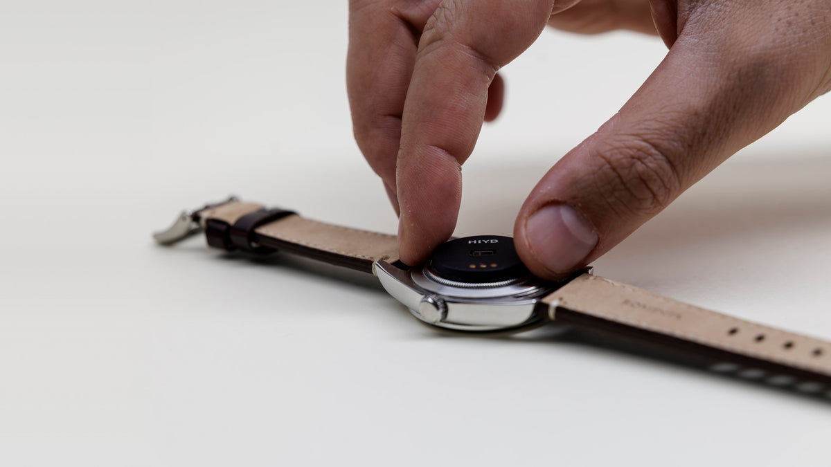 A hand pressing the HIYD sensor onto the back of a steel watch with a leather strap